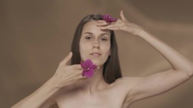 Ellerinde orkide çiçekleri olan bir kadın yüzüne dokunuyor, pürüzsüz ve yumuşak teninin tadını çıkarıyor. Stüdyodaki kahverengi arka planda bir kadının portresi. Güzellik konsepti, kozmetoloji