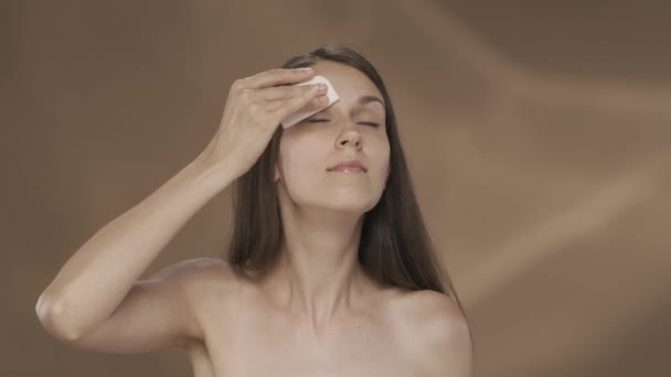 女性は顔を浄化するために綿パッドを使用します 茶色の背景にあるスタジオの若いセミナーの女性の肖像画 化粧品 ケアのコンセプト スローモーションHdr Bt2020 Hlg材料 — ストック動画