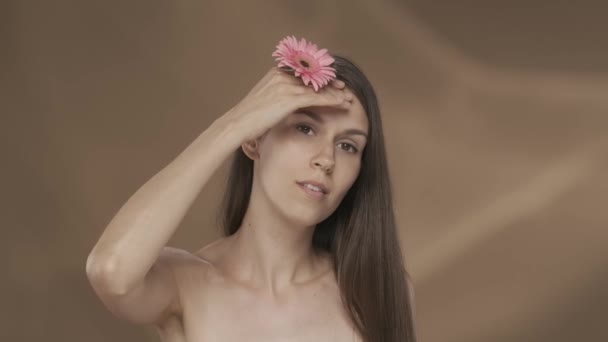 女性は彼女の顔 肩の皮膚にピンクの細菌の花で彼女の手に触れる 若いセミナーの女性は 茶色の背景にあるスタジオで彼女の完璧な肌を示しています コンセプト — ストック動画