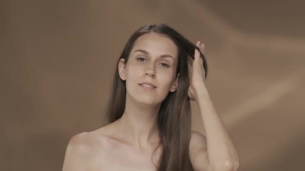 一个女人秀发柔滑 用手抚摩着它 带着棕色背景的年轻女子进入演播室 角蛋白矫直 美容美发的概念 — 图库视频影像