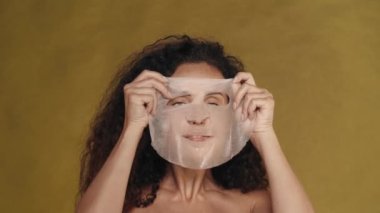 Bir kadın yüzüne kozmetik beyaz bir maske takar ve onu düzeltir. Stüdyoda sarı arka planda bir kadının portresi. Güzellik, kozmetoloji, özen kavramı