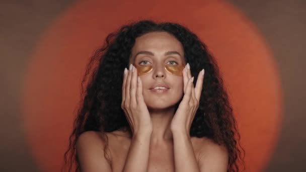 女性は彼女の目の下の黄金のパッチを滑らかにします スタジオで円形の光でオレンジ色の背景で化粧手続きをしているセミノードの女性 ビューティー — ストック動画
