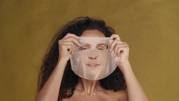 女性は彼女の顔に化粧品の白いシートマスクを適用し 滑らかにします 黄色の背景にあるスタジオのセミナーの女性の肖像画 ケアのコンセプト — ストック動画