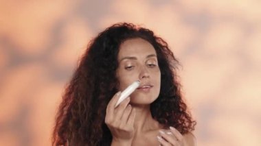 Bir kadın gözlerinin etrafındaki deri için jel kaldırır. Stüdyodaki uzun saçlı kıvırcık saçlı kadın turuncu arka planda ve yakın çekimde. Güzellik, kozmetoloji, cilt bakımı, spa