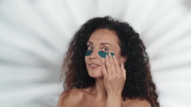 Bir kadın mavi hidrojel bantlarını gözlerinin altında düzeltip düzleştiriyor. Stüdyodaki gri arka planda röfleli bir kadın. Güzellik, kozmetoloji, cilt bakımı
