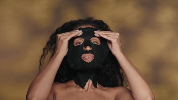 女性は顔から化粧品の黒いシートマスクを取り除きます 女性は肌に触れ 化粧効果を楽しんでいます ハイライト付きの黄色の背景にあるスタジオのセミノードの女性 コンセプト — ストック動画