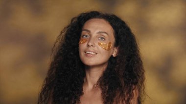 Uzun saçlı kıvırcık saçlı esmer kadın kozmetik ameliyatlar yapıyor. Stüdyoda, sarı arka planda, gözlerinin altında altın bantlı, yakın plan röfleli bir kadın portresi. Konsept