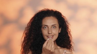 Bir kadın dudaklarıyla ilgilenir, dudak kremi sürer. Stüdyoda turuncu arka planda uzun saçlı kıvırcık saçlı esmer. Makyaj ve kozmetik, dudak derisi konsepti