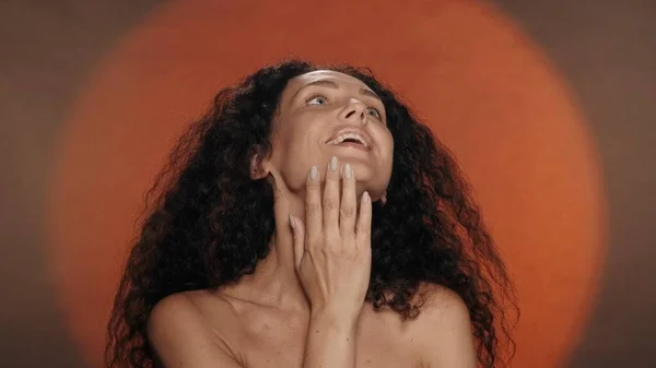 女性は肌の柔らかさと静けさを楽しみ 首に触れます 円形の光でオレンジ色のバックグラウンドにスタジオでセミナーカーリー女性の肖像画 美のコンセプト — ストック写真