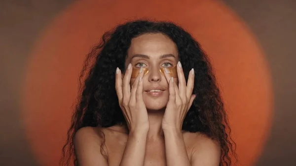 女性は彼女の目の下の黄金のパッチを滑らかにします スタジオで円形の光でオレンジ色の背景で化粧手続きをしているセミノードの女性 ビューティー — ストック写真