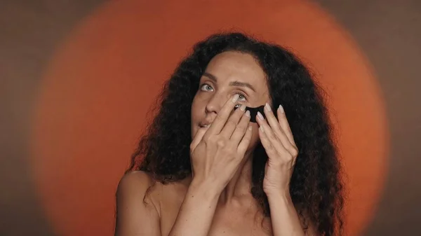 女性は目の下の皮膚に黒いヒドロゲルパッチを塗布する スタジオで円形の光でオレンジ色の背景で化粧手続きをしているセミノードの女性 ビューティー — ストック写真