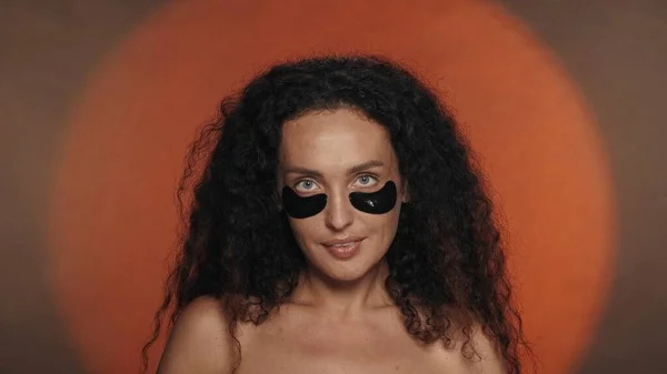長髪のカーリーブルネットの女性は 化粧品の手順を実行します スタジオのオレンジ色の背景に黒いパッチを被ったセミナーの女性の肖像画 イギリス — ストック写真