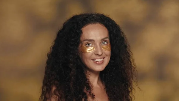 長髪のカーリーブルネットの女性は 化粧品の手順を実行します 黄色い背景のスタジオで彼女の目の下に金色のパッチを持つセミナーの女性の肖像画は ハイライトが近づいています コンセプト — ストック写真