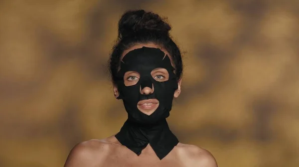 黒い化粧品のシートのマスクの女性と彼女の頭の上に束 ハイライト付きの黄色の背景にスタジオで化粧手順を実行するセミノードの女性 美のコンセプト — ストック写真