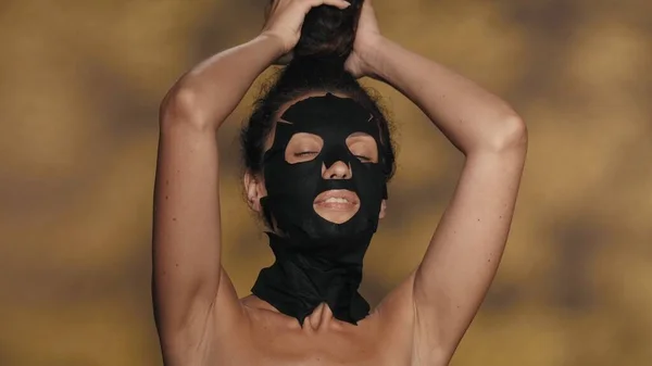黒い化粧品のシートのマスクの女性と彼女の頭の上に束 ハイライト付きの黄色の背景にスタジオで化粧手順を実行するセミノードの女性 美のコンセプト — ストック写真