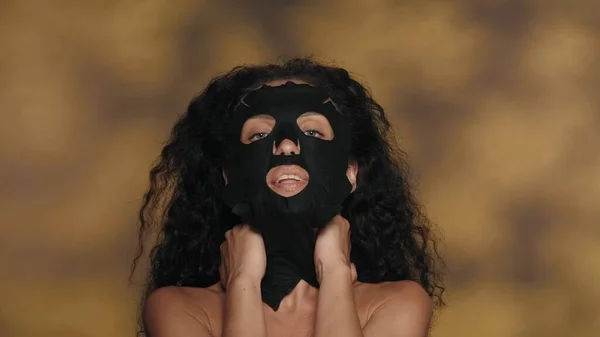 黒い化粧用シートのマスクをした女性 ハイライト付きの黄色の背景にスタジオで化粧手順を実行するセミノードの女性 ケアのコンセプト — ストック写真