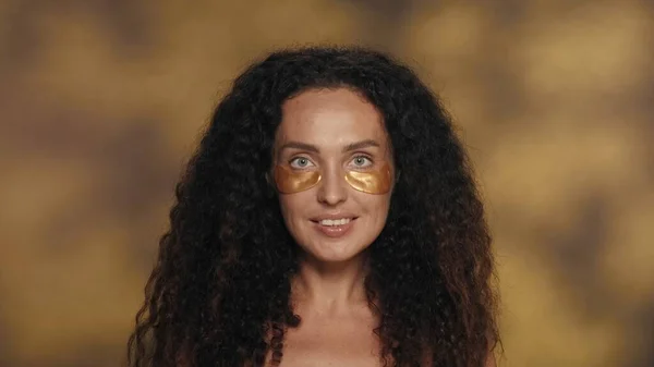 長髪のカーリーブルネットの女性は 化粧品の手順を実行します 黄色い背景のスタジオで彼女の目の下に金色のパッチを持つセミナーの女性の肖像画は ハイライトが近づいています コンセプト — ストック写真