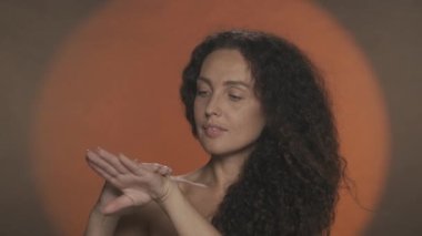 El bakımı ve koruma. Bir kadın ellerine cilt bakım kremi sürüp nemlendiriyor. Stüdyoda, turuncu arka planda dairesel ışıklı kıvırcık bir kadının portresi. Konsept: