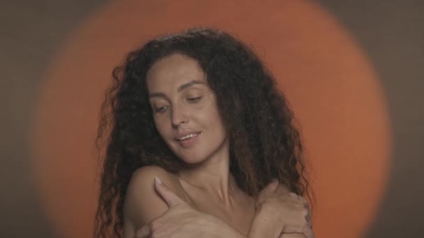 女人喜欢柔软柔滑的皮肤 摸着裸露的肩膀和胸部 画室里一个发亮的卷曲女人的肖像 背景是橙色的 有圆形的光 — 图库视频影像