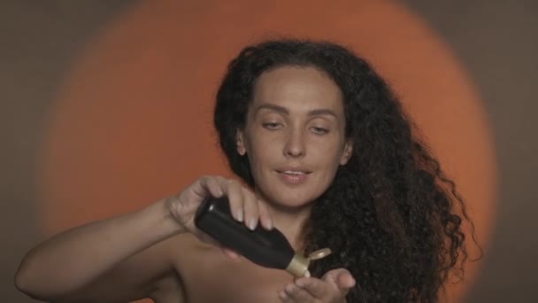 ハンドケアと保護 女性は彼女の手の黒いボトルからクリームを塗り 肌を保湿します 円形の光でオレンジ色の背景にスタジオでセミナーキュリーな女性の肖像画 — ストック動画