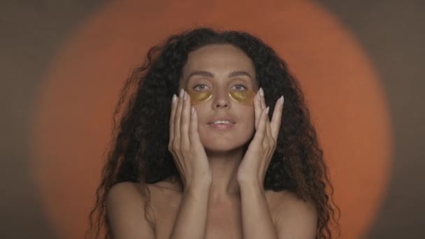 女性は彼女の目の下の黄金のパッチを滑らかにします スタジオで円形の光でオレンジ色の背景で化粧手続きをしているセミノードの女性 ビューティー ケアHdr Bt2020 Hlg — ストック動画