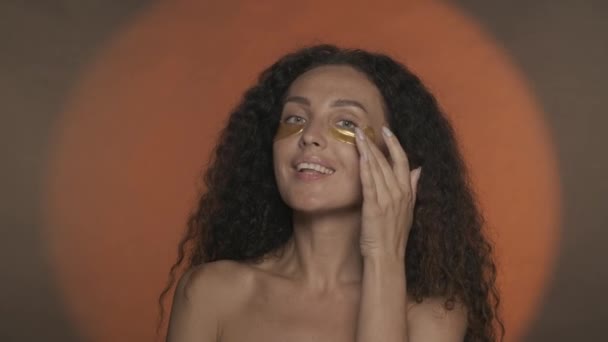 女性は彼女の目の下の黄金のパッチを滑らかにし 彼女の顔に触れることによって彼女の完璧な肌を楽しんでいます 円形のオレンジ色の背景でスタジオで化粧手続きを行うセミノードの女性 — ストック動画