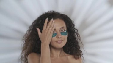 Bir kadın kozmetik bir işlem yapar ve pürüzsüz bir yüz derisi olduğunu gösterir. Stüdyoda mavi bantlı, gri arka planlı, parlak saçlı bir kadının portresi.