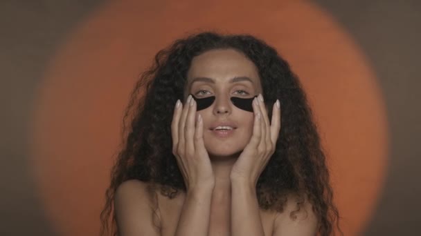 女性は目の下の黒いヒドロゲルパッチを滑らかにし 彼女の顔に触れることによって彼女の完璧な肌を楽しむ オレンジ色の背景でスタジオで化粧手続きを行うセミノードの女性 — ストック動画