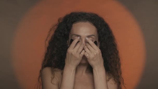 女性は目の下の黒いヒドロゲルパッチを取り除き 彼女の顔に触れることによって彼女の完璧な肌を楽しむ オレンジ色の背景でスタジオで化粧手続きを行うセミノードの女性 — ストック動画
