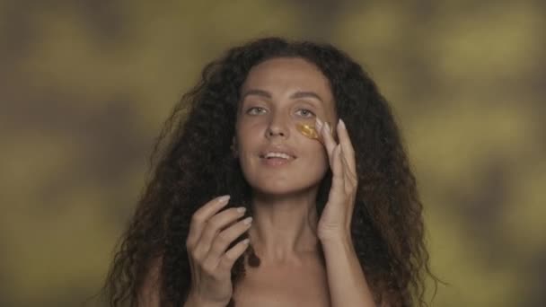 長髪のカーリーブルネットの女性は 化粧手順を実行し 彼女の目の下の皮膚に黄金のパッチを適用します ハイライトが近い黄色の背景にあるスタジオでセミナーの女性の肖像画 — ストック動画