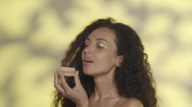 Stüdyoda elinde toz dolu bir kavanozla sarı bir arka planda parlak ışıklı güzel bir seminer kadını. Bir kadın pudraya üfledi. Makyaj yapıyorum. Güzellik konsepti, kozmetoloji