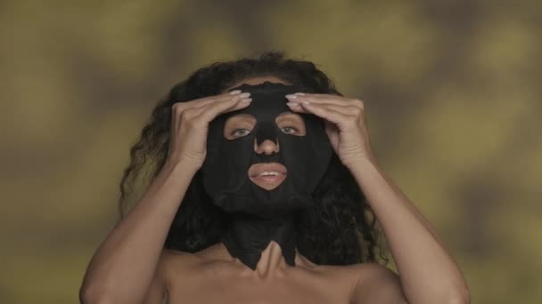 一个女人从她的脸上摘下一个化妆品的黑布面具 女人触摸皮肤 享受化妆品的效果 在带有亮点的黄色背景下 在演播室里表现突出的女人 — 图库视频影像
