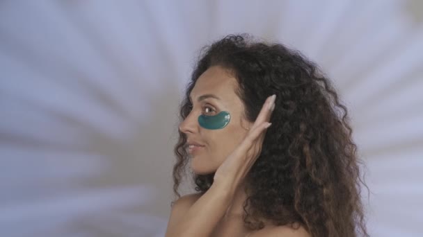 女性のプロフィール肖像画は 彼女の顔から彼女の手を取り除き ハイライト付きの青い背景のスタジオで開きます 女性は彼女の下に化粧品の手順 青いハイドロゲルパッチを実行します — ストック動画