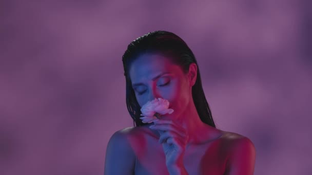 若い女性は近い視点で顔と肩をしています 彼女はピンクのバラの匂いを嗅いで カメラを見ている ピンクとブルーのカラーのコントラスト レジャーや製品の広告 — ストック動画