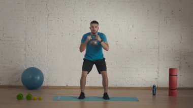 Evde kişisel spor eğitimleri. Atletik erkek fitness koçu çevrimiçi dersler için ev stüdyosunda egzersiz yapıyor. Spor kıyafetli bir adam kettlebell ile çömeliyor. Sağlık hizmetleri kavramı.