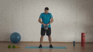 Evde kişisel spor eğitimleri. Atletik erkek fitness koçu çevrimiçi dersler için ev stüdyosunda egzersiz yapıyor. Spor kıyafetli bir adam kettlebell ile göğsüne vuruyor. Sağlık hizmetleri kavramı.