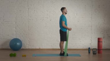 Yan görüş. Atletik erkek fitness koçu çevrimiçi dersler için ev stüdyosunda egzersiz yapıyor. Spor kıyafetli bir adam lastik bantla biceps egzersizi yapıyor. Sağlık hizmetleri kavramı.