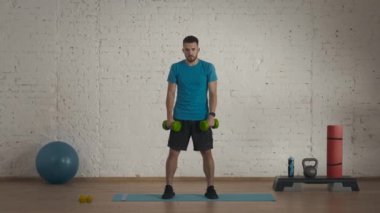 Atletik erkek fitness koçu çevrimiçi dersler için ev stüdyosunda egzersiz yapıyor. Spor kıyafetli bir adam yarım pozisyonda halterlerle salıncak sallıyor. Sağlık hizmetleri kavramı.