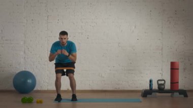 Atletik erkek fitness koçu çevrimiçi dersler için ev stüdyosunda egzersiz yapıyor. Spor giyimli bir adam lastik bant egzersiziyle yarım yamalak yan basamaklar yapıyor. Sağlık hizmetleri kavramı.