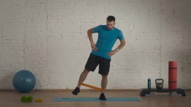 Atletik erkek fitness koçu çevrimiçi dersler için ev stüdyosunda egzersiz yapıyor. Spor kıyafetli bir adam lastik bantla yan bacak sallıyor. Sağlık hizmetleri kavramı.