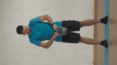 Dikey video. Evde kişisel spor eğitimleri. Atletik erkek fitness koçu çevrimiçi dersler için ev stüdyosunda egzersiz yapıyor. Spor kıyafetli bir adam kettlebell ile göğsüne vuruyor..
