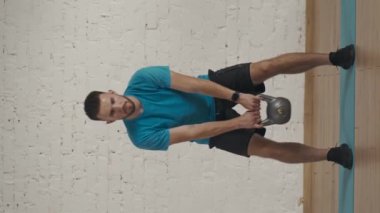 Dikey video. Evde kişisel spor eğitimleri. Atletik erkek fitness koçu çevrimiçi dersler için ev stüdyosunda egzersiz yapıyor. Spor kıyafetli bir adam elinde kettlebell ile çömeliyor..