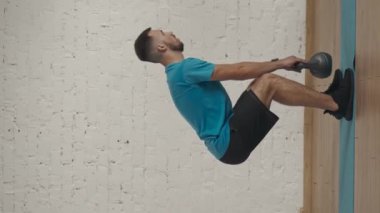 Dikey video. Kişisel eğitimler. Atletik erkek fitness koçu çevrimiçi dersler için ev stüdyosunda egzersiz yapıyor. Spor kıyafetli bir adam düz kollarla çömelmiş kettlebell yapıyor. Yan görünüm.