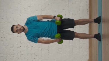 Dikey video. Evde kişisel spor eğitimleri. Atletik erkek fitness koçu çevrimiçi dersler için ev stüdyosunda egzersiz yapıyor. Spor kıyafetli bir adam halterli kol kaldırma egzersizi yapıyor..