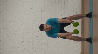 Dikey video. Atletik erkek fitness koçu çevrimiçi dersler için ev stüdyosunda egzersiz yapıyor. Spor kıyafetli bir adam yarım pozisyonda halterlerle salıncak sallıyor. Ön ve yan görünüm.