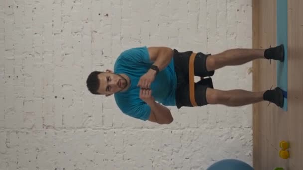 垂直录像 运动健美教练在家庭工作室为在线课程做运动 身穿运动服的男子用橡皮筋作了半弯腰的侧步运动 保健概念 — 图库视频影像