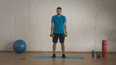 Kapalı alanda spor eğitimi. Atletik erkek fitness koçu çevrimiçi dersler için ev stüdyosunda egzersiz yapıyor. Spor kıyafetli bir adam yeşil halterlerle egzersiz yapıyor..