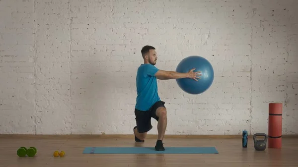 Athletic male fitness coach doing exercises at the home studio for online classes. Man in sportswear doing lunges exercise holding a fitball.