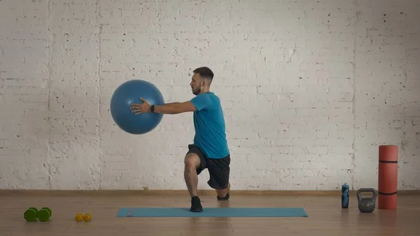 Sport indoors. Athletic man fitness coach doing exercises at the home studio for online classes. Man in sportswear doing lunges exercise holding a fitball.
