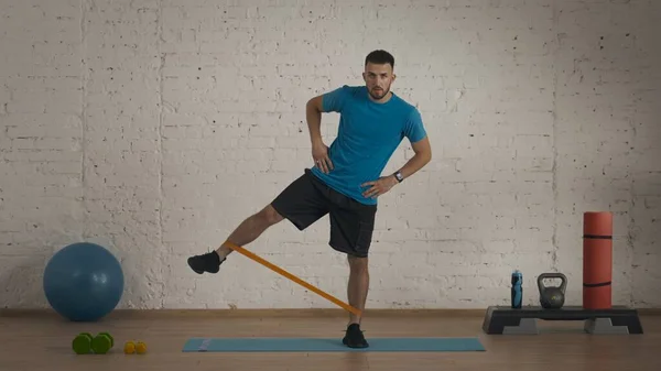 Athletic male fitness coach doing exercises at the home studio for online classes. Man in sportswear doing side leg swing with elastic band. Sport indoors.